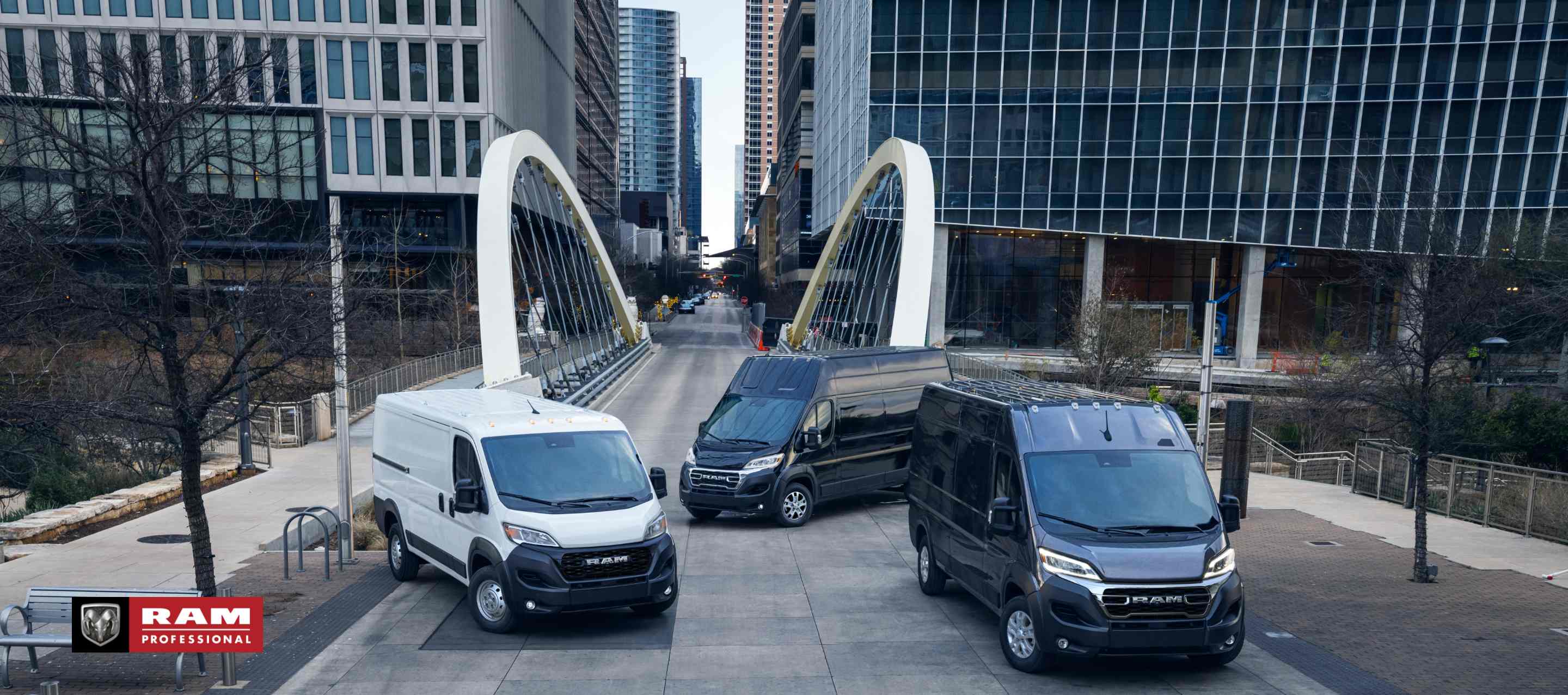 Three 2024 Ram ProMaster models, from left to right: a white Ram ProMaster 1500 Tradesman Standard Roof Cargo Van, a black Ram ProMaster 3500 SLT+ Super High Roof Cargo Van and a black Ram ProMaster 3500 SLT High Roof Cargo Van parked just beyond a bridge in the center of a busy large city. Ram Professional.