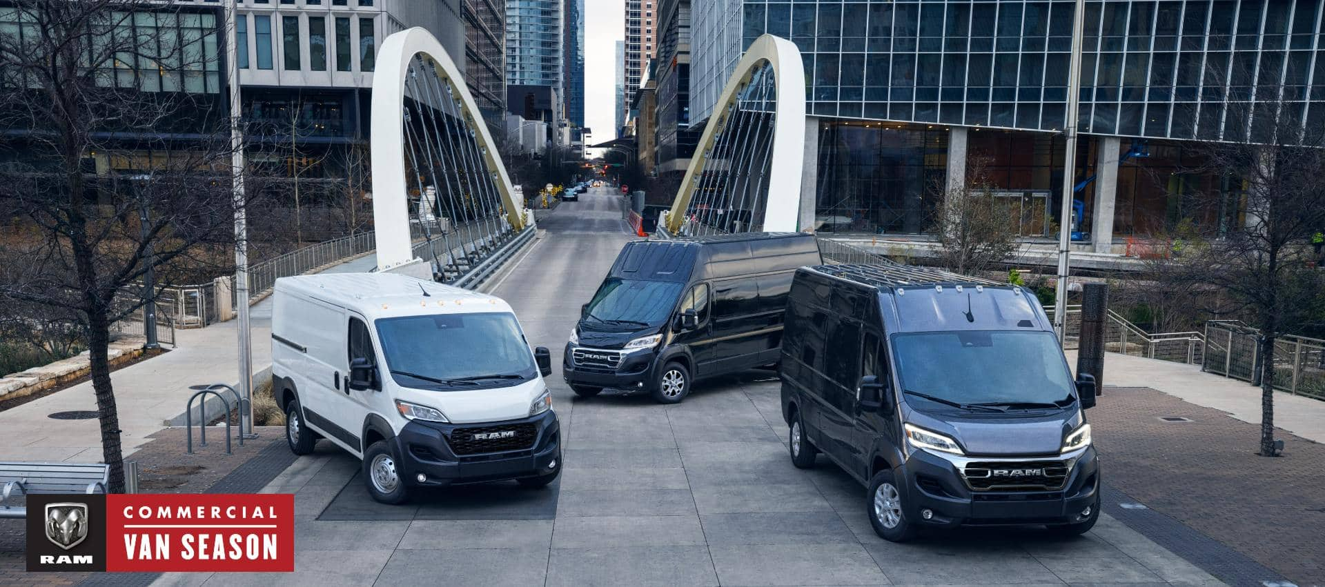 Three 2024 Ram ProMaster models, from left to right: a white Ram ProMaster 1500 Tradesman Standard Roof Cargo Van, a black Ram ProMaster 3500 SLT+ Super High Roof Cargo Van and a black Ram ProMaster 3500 SLT High Roof Cargo Van parked just beyond a bridge in the center of a busy large city. Commercial Van Season.