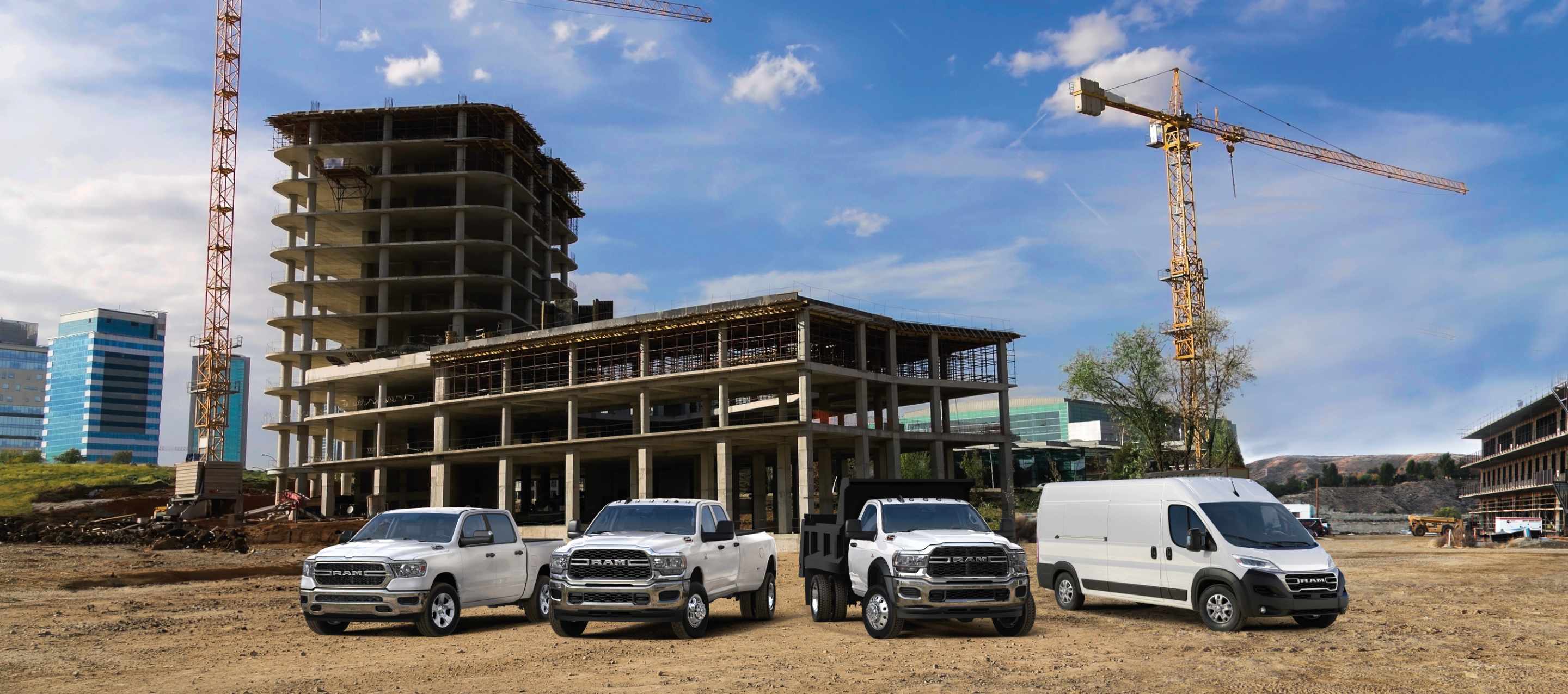 A Ram Brand lineup of four vehicles all in white, parked side-by-side at a construction site. From left to right: a 2024 Ram 1500 Tradesman 4x4 Crew Cab, a 2024 Ram 3500 Tradesman 4x4 Crew Cab, a 2024 Ram 5500 Tradesman Chassis Cab Regular Cab 4x4 with dump body upfit and a 2024 Ram ProMaster 3500 SLT High Roof Cargo Van.