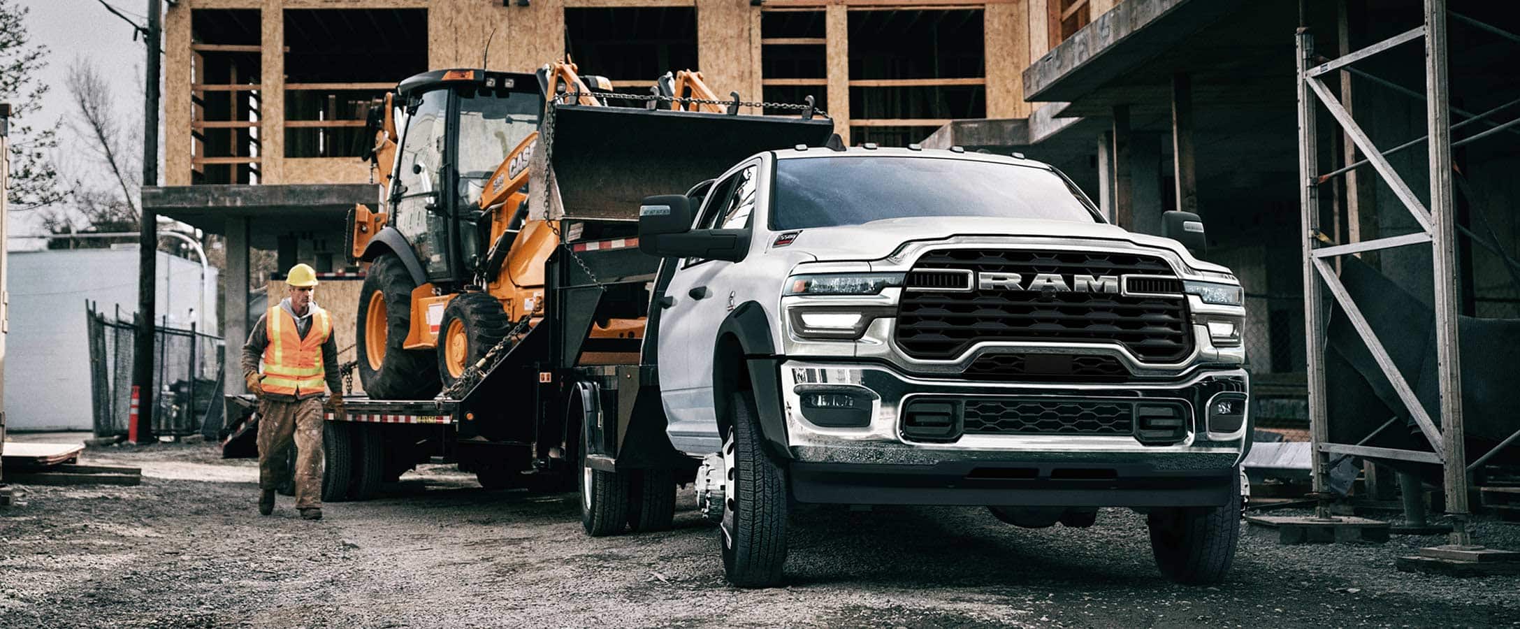A white 2025 Ram 5500 Big Horn Chassis Crew Cab on a construction site with an excavator in tow.